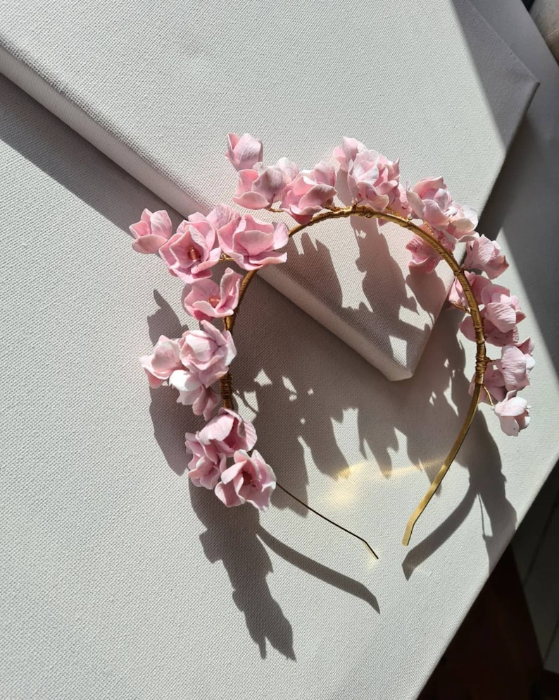 May include: A pink flower crown headband with a gold wire base. The headband is made of delicate pink flowers and is perfect for a special occasion.