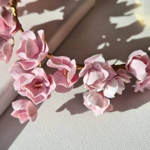 May include: A delicate pink floral crown made of artificial flowers, with a gold wire base. The crown is lying on a white surface.