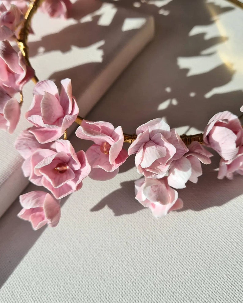 May include: A delicate pink floral crown made of artificial flowers, with a gold wire base. The crown is lying on a white surface.