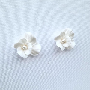 May include: Two white floral earrings with a pearl center are displayed against a white background. The earrings are shaped like delicate flowers, with multiple petals and a small, round pearl in the center of each. The earrings are likely made of porcelain or a similar material.