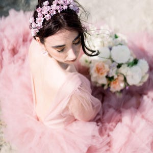 May include: A woman wearing a pink tulle dress and a floral crown. She is holding a bouquet of white, pink, and peach flowers.