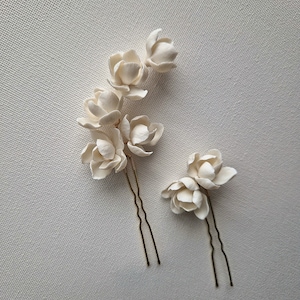 May include: Two white ceramic flower hair pins with gold metal stems. The flowers are arranged in a cluster of three and two.