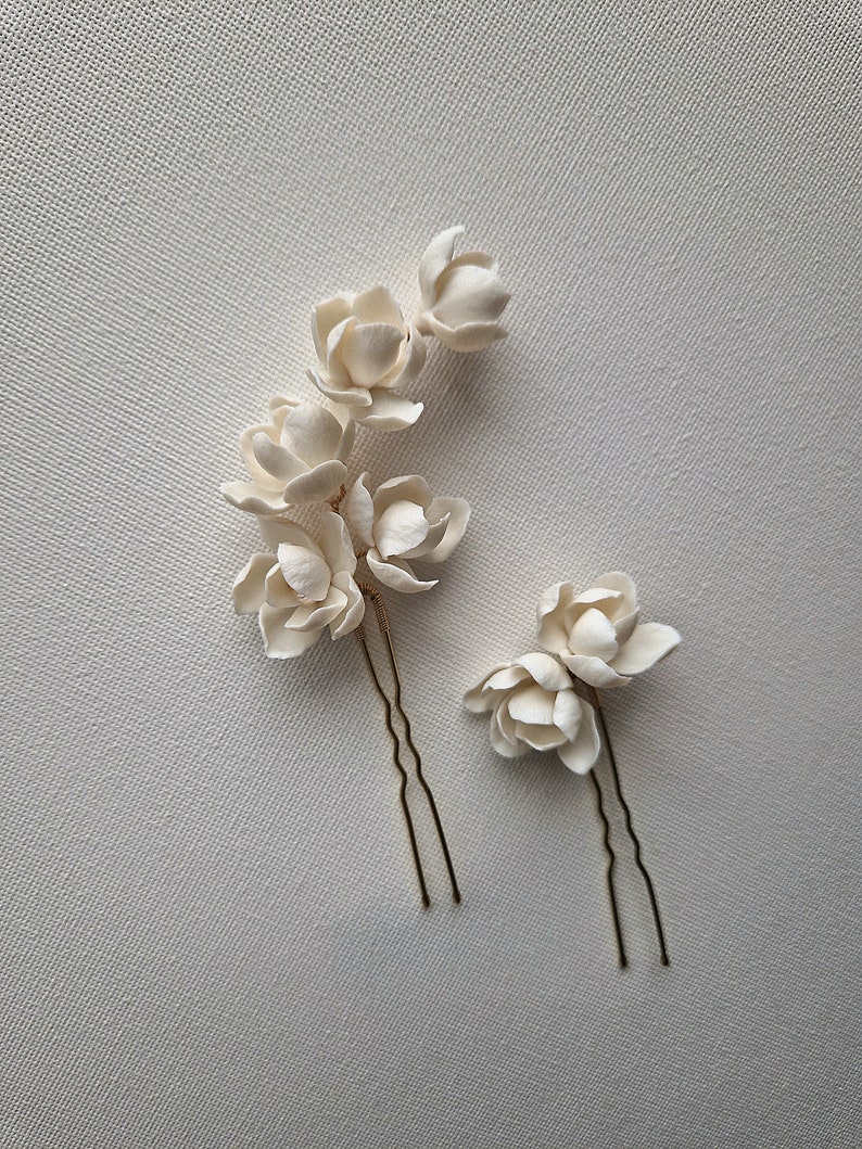 May include: Two white ceramic flower hair pins with gold metal stems. The flowers are arranged in a cluster of three and two.