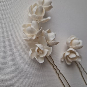 May include: Two white porcelain flower hair pins with gold wire stems. The larger pin has five flowers and a pearl accent. The smaller pin has two flowers.