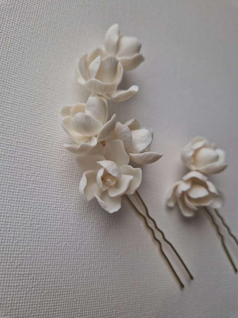 May include: Two white porcelain flower hair pins with gold wire stems. The larger pin has five flowers and a pearl accent. The smaller pin has two flowers.