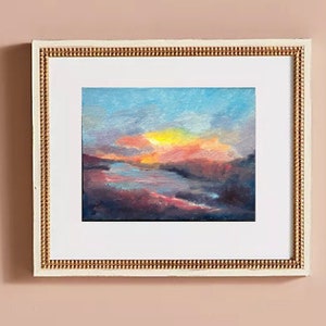 Puede incluir: Una pintura enmarcada de una puesta de sol sobre un lago. El cielo es una mezcla de azul, rosa y naranja, con un sol amarillo brillante en el centro. El lago es de un azul oscuro, con algunos árboles y arbustos en la orilla.