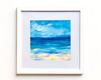 May include: A framed abstract painting of a seascape. The artwork features a bright blue sky with white clouds above a deep blue ocean. The foreground shows a colorful beach with pink, yellow, and green hues. The painting is in a white frame.