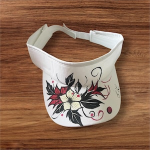 May include: White visor with a floral design. The visor features a white brim and adjustable strap. The floral design includes red, black, and yellow flowers with black leaves and red accents. The visor is made of a canvas-like material.