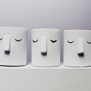 Può includere: Tre vasi in ceramica bianca con semplici occhi e nasi dipinti di nero. I vasi sono disposti in fila su una superficie bianca.