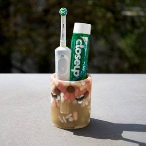 May include: A decorative toothbrush holder with a face design, holding an electric toothbrush and a tube of Closeup toothpaste. The holder is beige with pink and white accents. The toothpaste tube is green and white.