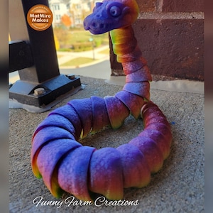 May include: A rainbow-colored, segmented snake toy made of plastic. The snake is curled into a circle with its head raised. The toy is made by Funny Farm Creations.