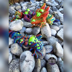 May include: A pair of colorful, 3D printed, dragon-like figures with green, blue, and red accents. The figures are posed on a bed of gray rocks.