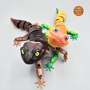 May include: Two articulated toy lizards, one black and brown with yellow eyes, the other orange and yellow with blue eyes. The lizards are posed as if they are playing together. The background is a light gray.