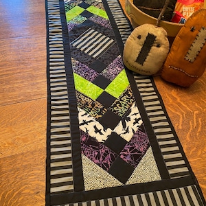 May include: A rectangular quilted table runner featuring a central chevron pattern in black, purple, and lime green fabrics with prints of crows, spiderwebs, and the text "SCARY TREATS". The runner is bordered by black and grey striped fabric.
