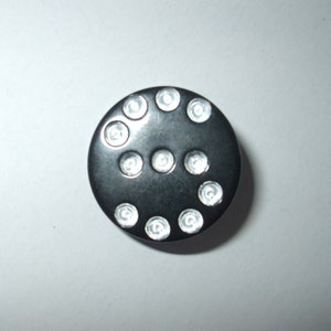 May include: A black button with a circle of silver-colored dots around the edge.