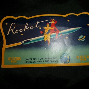 May include: Vintage packaging for Rocket brand needles. The packaging features a blue and green background with a silver needle flying through space with two figures riding on it. The text on the packaging reads "Rocket", "Silver Eye", "Contains 100 Assorted Needles and 1 Threader", and "Gold Eye".