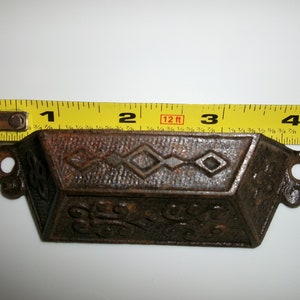 May include: A rusty cast iron drawer pull with a diamond pattern and ornate design. The pull measures approximately 2 1/2 inches wide.
