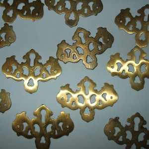 May include: A collection of ornate, brass-colored keyhole escutcheons. Each escutcheon features a decorative, symmetrical design with a keyhole and multiple cut-out shapes. The hardware is likely for furniture or decorative purposes.