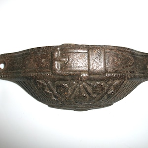 May include: A cast iron drawer pull with a decorative design featuring a belt buckle. The pull is brown and has a slightly distressed finish.