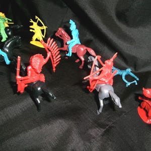 May include: A collection of plastic toy Native American figures on horseback. The figures are in various colors, including red, blue, green, and yellow. The figures are holding weapons and riding horses.