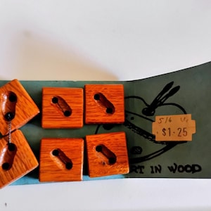 May include: Six square wooden buttons with a reddish-brown finish are displayed on a teal card. Each button has a unique carved design and two black holes for sewing. The card has the text "ART IN WOOD" and a price tag.