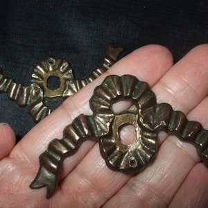 May include: Two antique brass ribbon-shaped drawer pulls with a decorative, ornate design. The pulls have a hole in the center for attaching to a drawer.