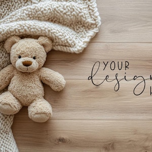 Pode incluir: Um urso de pelúcia bege claro repousa parcialmente sob um cobertor texturizado creme. O urso tem um nariz castanho e uma expressão amigável. As palavras "YOUR design HERE" estão escritas em itálico no fundo de madeira.