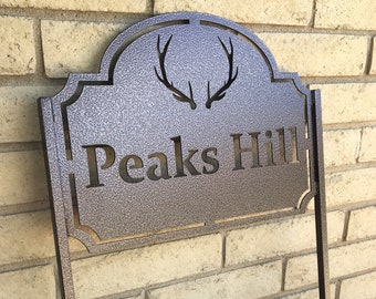 May include: A brown metal sign with a decorative arch and a pair of antlers. The sign says "Peaks Hill" in a stylized font.