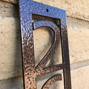 May include: A close-up of a copper-coloured metal house number sign with the number "2" in a modern font. The sign has a hammered texture and is mounted on a brick wall.