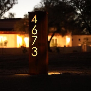 May include: A rectangular, dark brown address post with the numbers 4673 illuminated in a warm, golden light. The post is set in a grassy area, with a house and trees in the blurred background. The numbers are cut out of the metal.