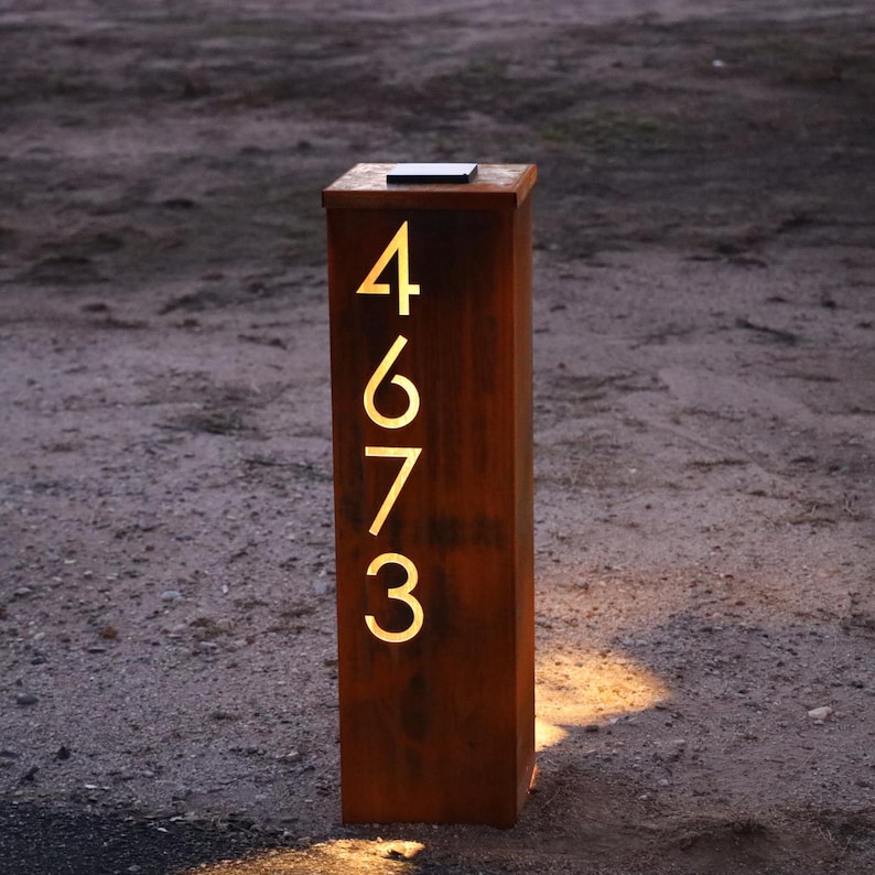 May include: A rectangular, brown metal address post with the numbers 4673 illuminated in a warm, yellow light. The post is standing upright on a sandy surface, with a small, dark panel on top.