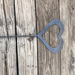 Pu&ograve; includere: Un ritaglio a forma di cuore in metallo attaccato a un lungo bastone di metallo. Il bastone e il cuore sono di colore grigio scuro. Il cuore &egrave; posizionato su una superficie di legno.