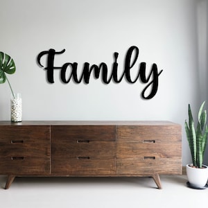 Peut inclure: Une pancarte en bois noir avec le mot "Family" en écriture cursive, fixée sur un mur blanc au-dessus d'une commode brun foncé à six tiroirs. Une plante verte est placée à gauche de la commode et une autre plante verte à droite.