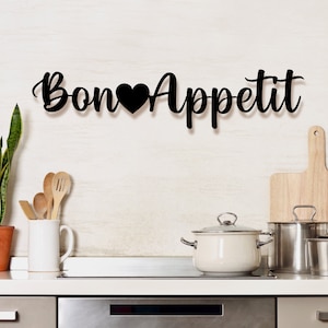 Puede incluir: Decoración de pared de metal negro con el texto "Bon Appetit" y un corazón entre las palabras. La decoración está colgada en una pared blanca de azulejos sobre una encimera de cocina con tablas de cortar de madera, ollas y utensilios.