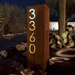May include: A rectangular, weathered metal address sign with the numbers "3360" illuminated in a warm, glowing light. The sign is set outdoors, with a desert landscape in the background.