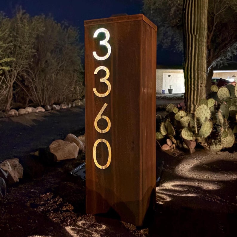 May include: A rectangular, weathered metal address sign with the numbers "3360" illuminated in a warm, glowing light. The sign is set outdoors, with a desert landscape in the background.