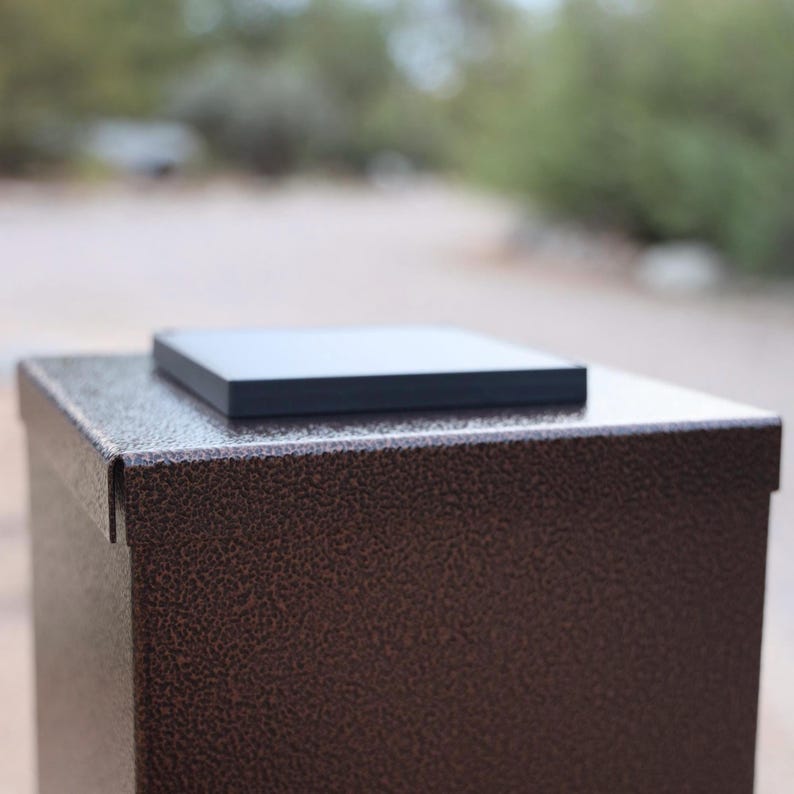 May include: A bronze-colored metal box with a textured finish, topped with a flat, black square. The box has a rectangular shape and appears to be made of durable material. The background is blurred.