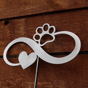 May include: A white metal infinity symbol with a paw print and a heart in the center. The symbol is attached to a metal rod.