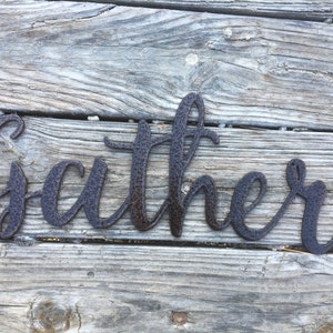 May include: A rustic metal sign with the word "Gather" in a cursive font. The sign is painted in a dark brown color and has a textured finish.