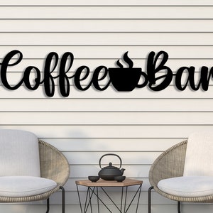 Può includere: Un cartello in metallo nero con le parole "Coffee Bar" in un font corsivo. Una tazza di caffè stilizzata con vapore è tra le parole. Il cartello è montato su una parete di listelli di legno bianco. Due sedie in vimini con cuscini bianchi sono di fronte al cartello.