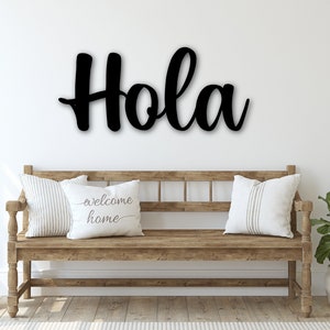 Op de afbeelding: Zwart houten bord met het woord "Hola" in een cursief schrift. Het bord hangt aan een witte muur boven een houten bank met kussens. De bank staat in een gang met een groene plant in een witte pot.