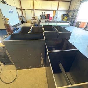May include: Five black metal planter boxes of various sizes are arranged in a workshop setting. The boxes are made of thick metal and have a simple, industrial design.