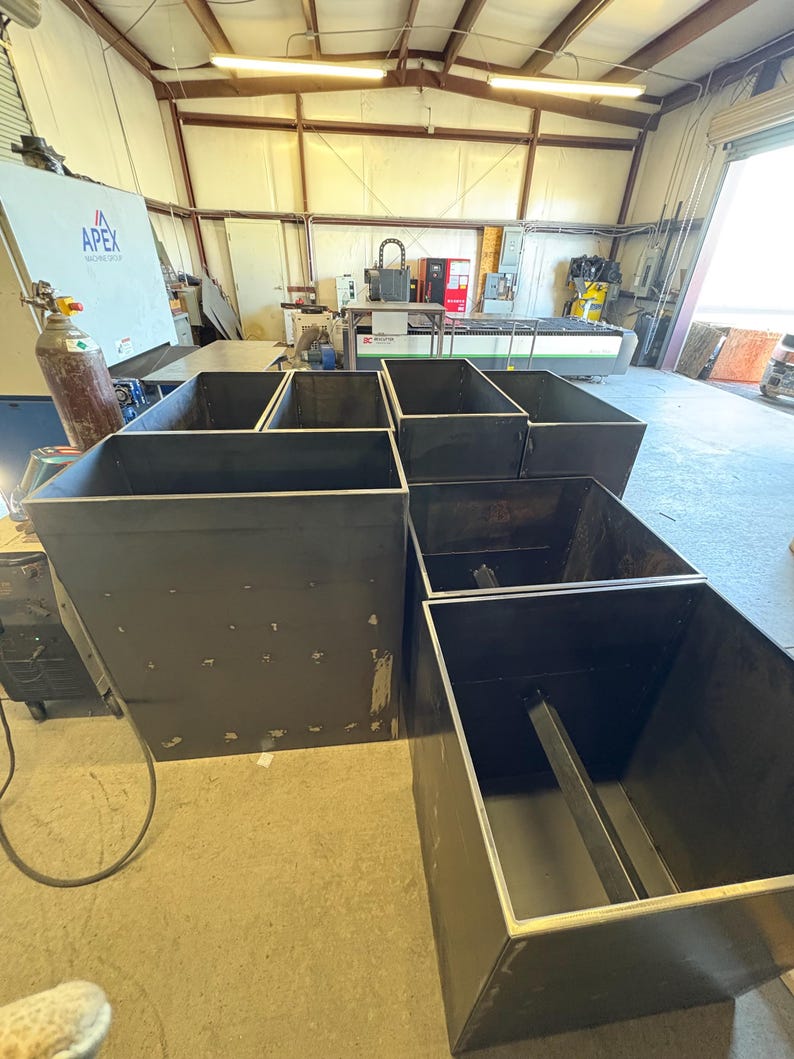 May include: Five black metal planter boxes of various sizes are arranged in a workshop setting. The boxes are made of thick metal and have a simple, industrial design.