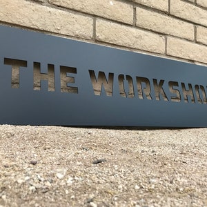 May include: A dark gray metal sign with the words "THE WORKSHOP" cut out. The sign is rectangular and mounted on a brick wall. The sign is angled slightly, and the ground is covered in small pebbles.