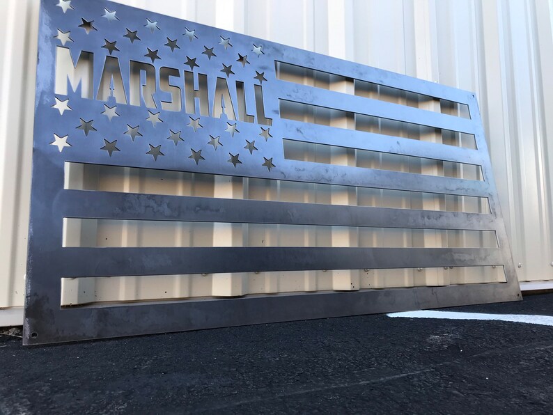 May include: A metal American flag with the word "MARSHALL" cut out of the stars. The flag is made of a dark metal and has a rustic look.