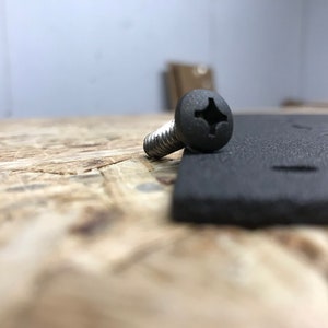 May include: A close-up of a black screw with a Phillips head, lying on a black surface. The screw is partially visible, with the head facing the camera.