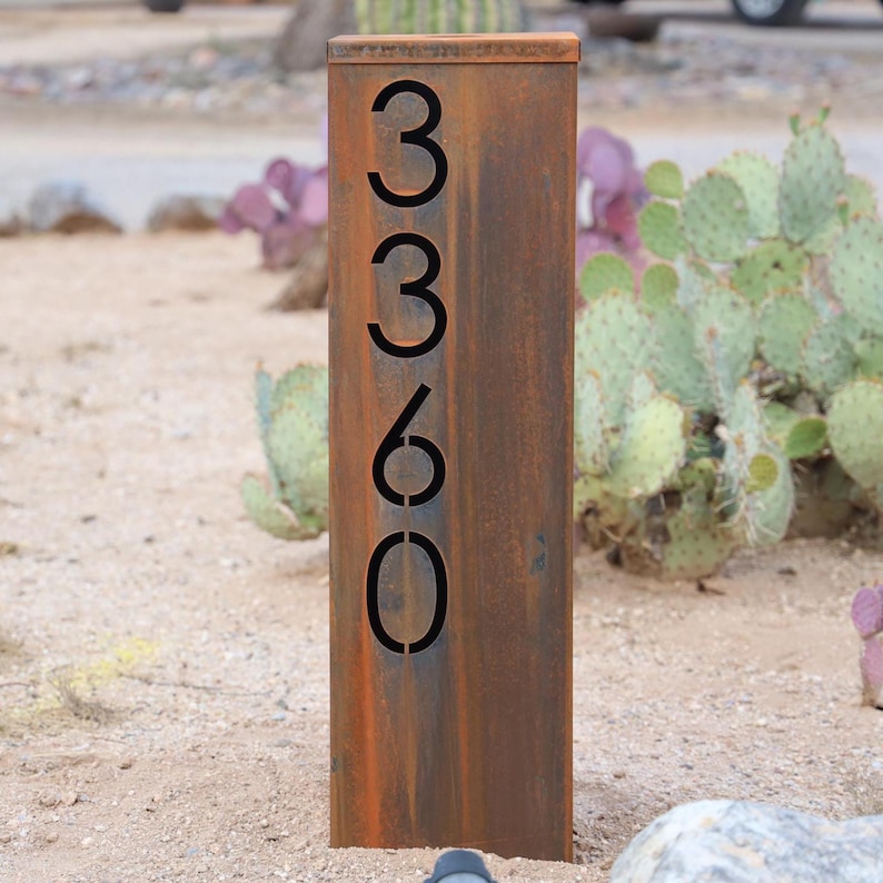 May include: A rectangular, weathered metal address sign with the numbers "3360" cut out. The sign is a rusty brown color and stands upright in a desert landscape with cacti and sand.