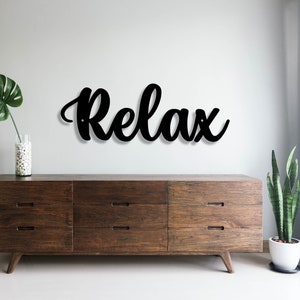 May include: Black wooden sign with the word "Relax" in a cursive font, hanging on a white wall above a dark brown dresser with six drawers. A green plant is in a white pot on the right side of the dresser.