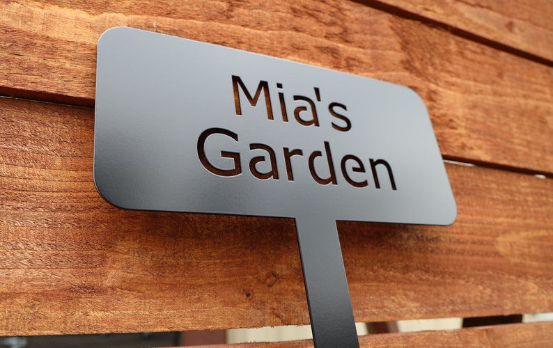 Puede incluir: Letrero de jard&iacute;n de metal negro con el texto "Mia's Garden" recortado.