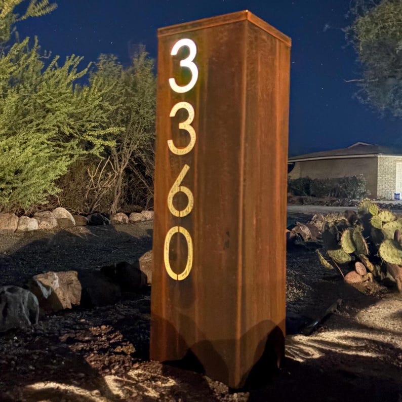 May include: A tall, rectangular address sign made of weathered, rusty metal. The numbers "3360" are illuminated in a warm, golden light. The sign is set outdoors, surrounded by dark foliage and a night sky.